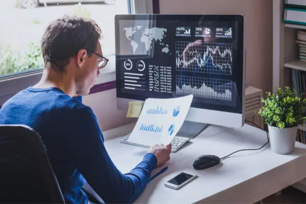 Man sitting in front of the computer screen with a dashboard on it, and looking into the printout of a diagram in his hand.